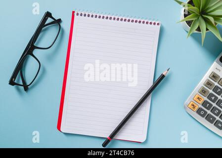 Top view of blank notebook, glasses, pen, calculator and green plant on blue office desk Stock Photo