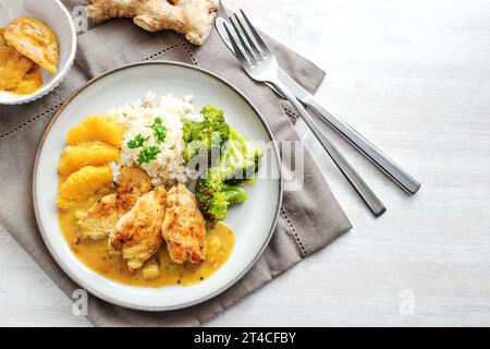Fried chicken fillet in orange ginger sauce with broccoli vegetables and rice, on a plate with cutlery and a gray napkin on a white table, top view fr Stock Photo
