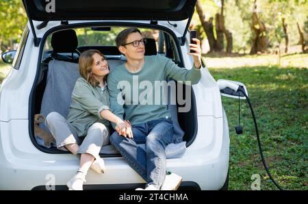 Loving couple with smartphones sits in an electric car's trunk. Stock Photo