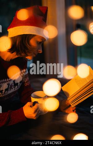Woman in Santa hat and Christmas sweater with glowing garland and book. Cozy Christmas evening. Girl with fairy tales book and electric garland. Stock Photo