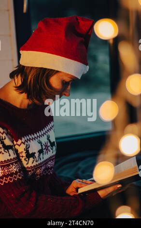 Woman in Santa hat and Christmas sweater with glowing garland and book. Cozy Christmas evening. Girl with fairy tales book and electric garland. Stock Photo