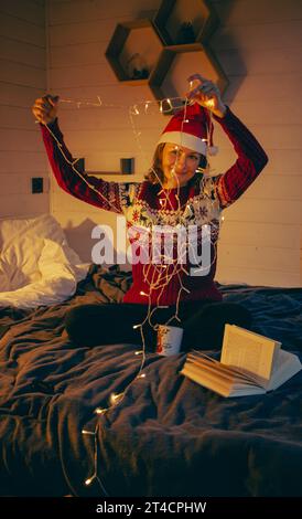 Woman in Santa hat and Christmas sweater with glowing garland and book. Cozy Christmas evening. Girl with fairy tales book and electric garland. Stock Photo