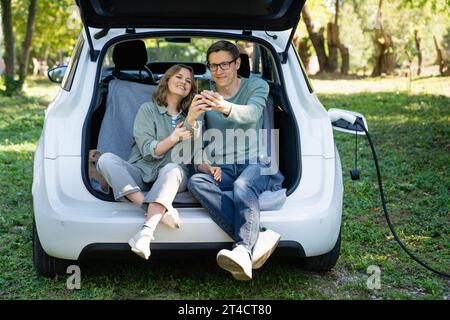 Loving couple with smartphones sits in an electric car's trunk. Stock Photo
