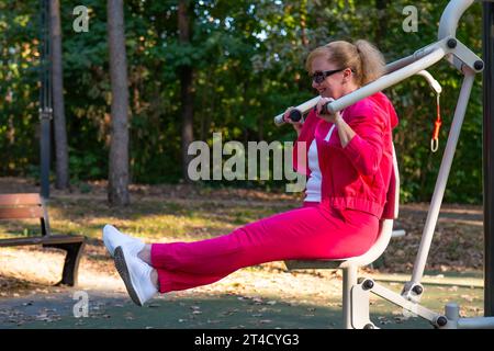 A middle-aged woman in a red tracksuit and glasses is engaged in an outdoor sports ground in the park. Stock Photo