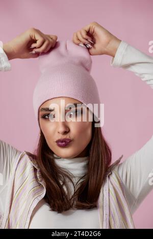 portrait of happy playful woman keeps hands on pink hat on pastel pink background. Stock Photo