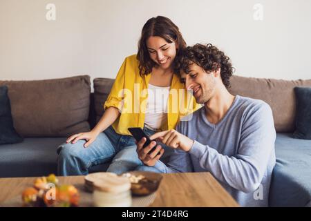 Portrait of young happy couple who is looking at smartphone at home. Stock Photo