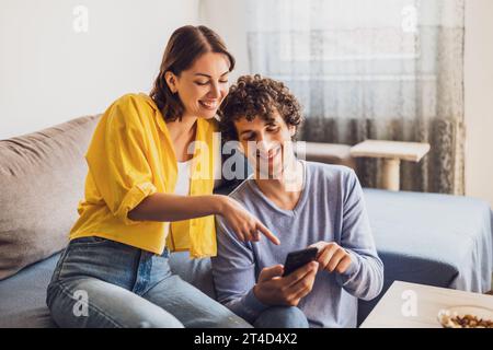 Portrait of young happy couple who is looking at smartphone at home. Stock Photo