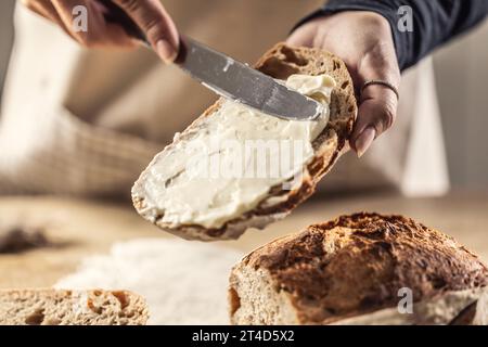 A woman makes delicious bread, spreads cream cheese with a cutlery knife - Close up. Woman hands spreading cream cheese on bread slice. Stock Photo