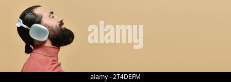 side view banner, pleased bearded man in turtleneck jumper listening music in headphones on beige Stock Photo