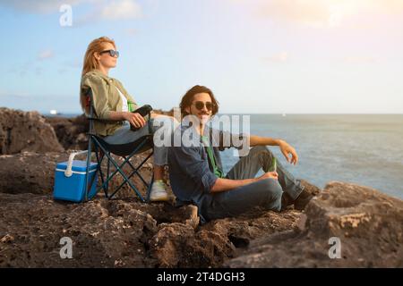Portrait Of Man And Woman Resting In Camping Chairs On Beach Rocks Stock Photo