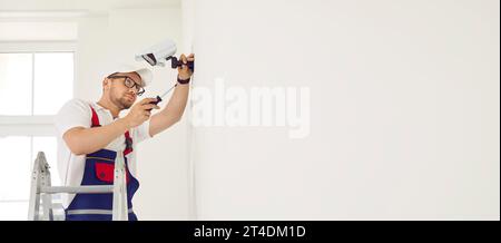 Male technician adjusts, installs or repairs modern CCTV camera on white wall in office or home. Stock Photo
