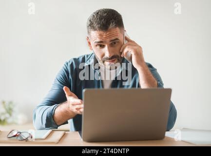 Displeased Man Talking On Cellphone Gesturing To Laptop At Workplace Stock Photo