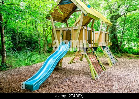 Colored green slide attached to roofed viewing platforms with climbing nets and grabs handles set in  a wooded area with lots of trees and shrubs Stock Photo