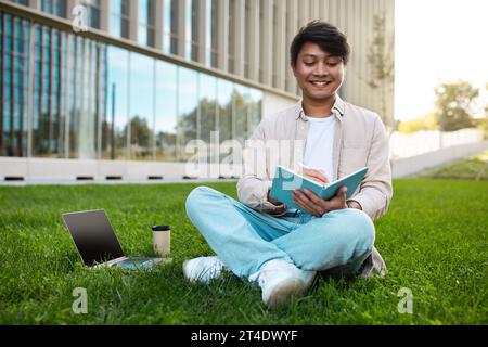 Happy young asian guy student with laptop sitting outdoors Stock Photo