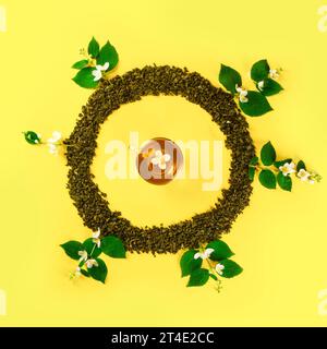Mockup of dried tea leaves and fresh jasmine flowers shaped in round Frame with tea cup in center isolated on plain yellow background Stock Photo