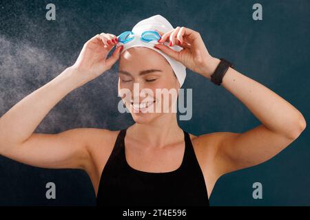 Portrait of a happy female swimmer adjusting goggles over a blue background Stock Photo