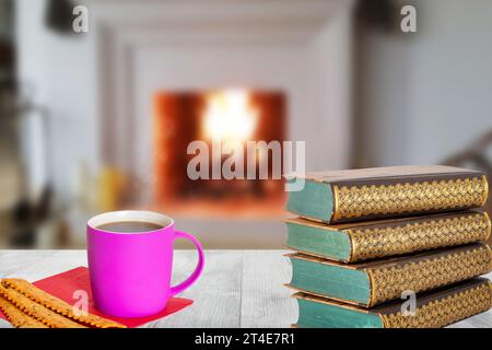 Winter christmas books background. A stack of antique books with a coffee mug and Italian pastries on a wooden table over abstract blurred fireplace. Stock Photo