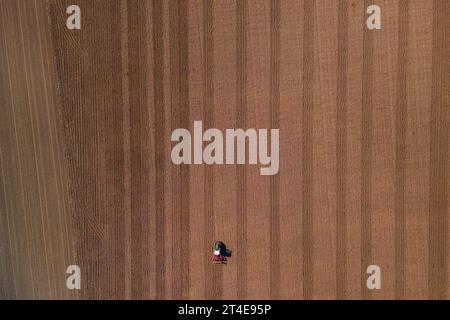 A small lonely tractor with plow on a large brown field with plowed earth seen directly from above Stock Photo