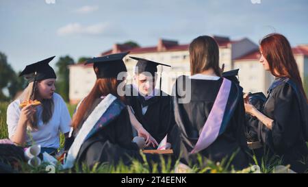 Graduates in black suits eating pizza in a city meadow. Stock Photo