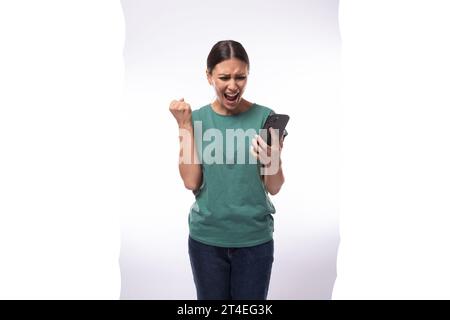 A 30 year old slender European brunette woman with a ponytail dressed in a green T-shirt and jeans screams joyfully while watching a video on the Stock Photo