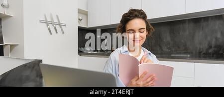 Portrait of brunette woman does homework, works from home, prepares for exam in kitchen, sits with notebook and laptop Stock Photo