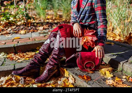 Close up of burgundy red leather knee high boots. Fashionable woman wearing stylish checkered blazer skirt sitting in fall park with purse among falle Stock Photo