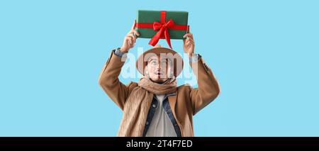 Stylish young man in winter clothes holding gift on light blue background Stock Photo