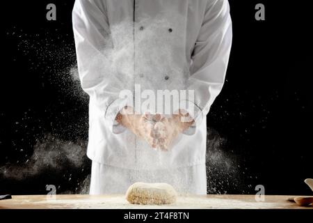 Young chef clapping and sprinkling flour over dough on table against black background Stock Photo