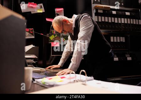 Inspector working overtime at criminal investigations case, analyzing evidence files in arhive room. Senior private detective checking criminology report, standing in police department Stock Photo