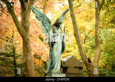 an antique angel figure with a palm branch stands on a grave of a cemetery in front of an autumnal colorful leafy forest in a blurred background Stock Photo
