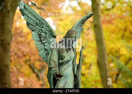 an antique angel figure with a palm branch stands on a grave of a cemetery in front of an autumnal colorful leafy forest in a blurred background Stock Photo