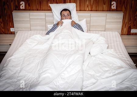 Frightened man in bed hiding under the covers Stock Photo