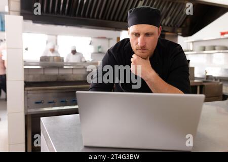 Serious caucasian male chef in black uniform thinking and working over laptop on counter in kitchen Stock Photo