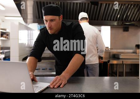 Caucasian male chef in black uniform working over laptop on counter with student in background Stock Photo