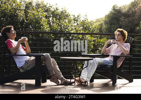 Happy diverse gay male couple sitting on chairs and drinking coffee at balcony Stock Photo