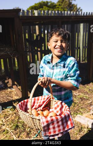 Portrait of happy biracial boy holding basket with eggs in front of henhouse Stock Photo