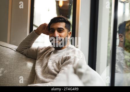 Pensive biracial man sitting on sofa and smiling in sunny living room Stock Photo