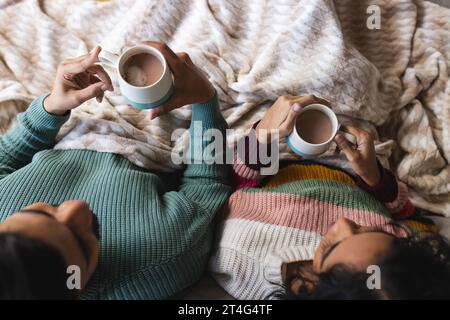 Biracial couple sitting on sofa under blanket and drinking cocoa in living room at home Stock Photo