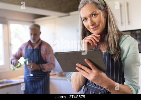 Portrait of happy senior caucasian couple cooking dinner, using tablet in kitchen Stock Photo