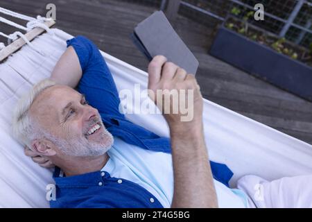 Happy senior caucasian man lying in hammock and using tablet Stock Photo