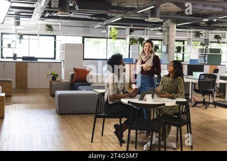 Happy diverse female colleagues talking at table with tablet in office Stock Photo