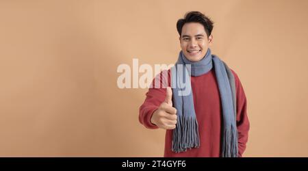 Happy and positive Asian man in red sweater and scarf giving thumbs up with ad empty space isolated on beige color background. Stock Photo