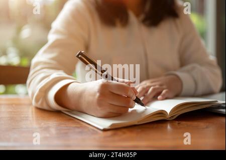Cropped image of an Asian woman in casual clothes writing something in her book, making lists, taking notes, or keeping her diary at a table indoors. Stock Photo