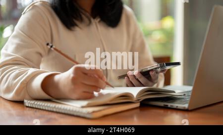 Cropped image of an Asian woman holding her smartphone while taking notes in a book, doing homework or studying an online class at a table indoors. Stock Photo