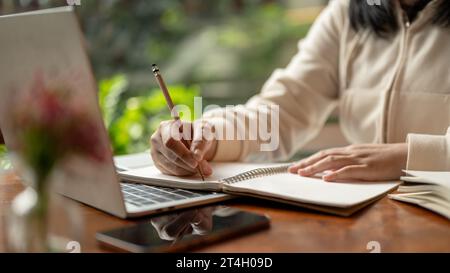 Cropped image of an Asian woman in casual clothes writing something in her book, taking notes while studying online, and working at a table indoors. Stock Photo