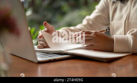 Cropped image of an Asian female student in casual clothes writing something in her book, taking notes while studying online, preparing for the exam, Stock Photo