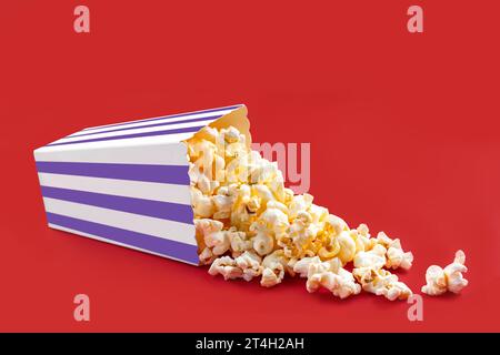 Tasty cheese popcorn falling out of a purple striped carton box or bucket, isolated on red background. Scattering of popcorn grains. Fast food, snack. Stock Photo