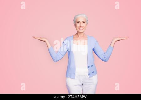 Beautiful nice cheerful smiling gray-haired old lady wearing casual, showing two options with palms. Isolated over pastel violet purple background Stock Photo