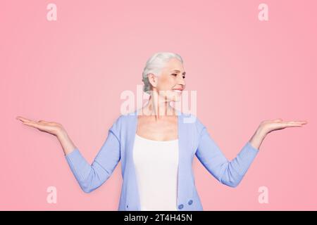 Beautiful nice cheerful smiling gray-haired old lady wearing casual, showing two options with palms, looking at one alternative. Isolated over pastel Stock Photo