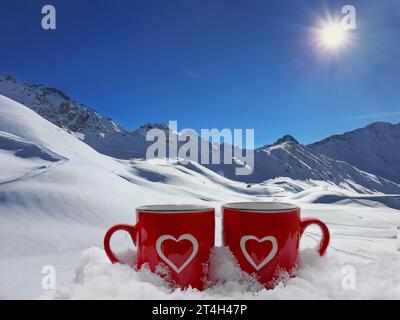 two red mug with a heart drawn on it in the snow in front of a snowy mountain landscape- winter sports Stock Photo